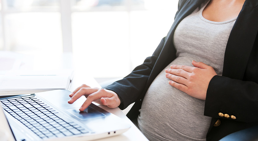 A woman sits at a desk while working on her laptop holding her pregnant belly